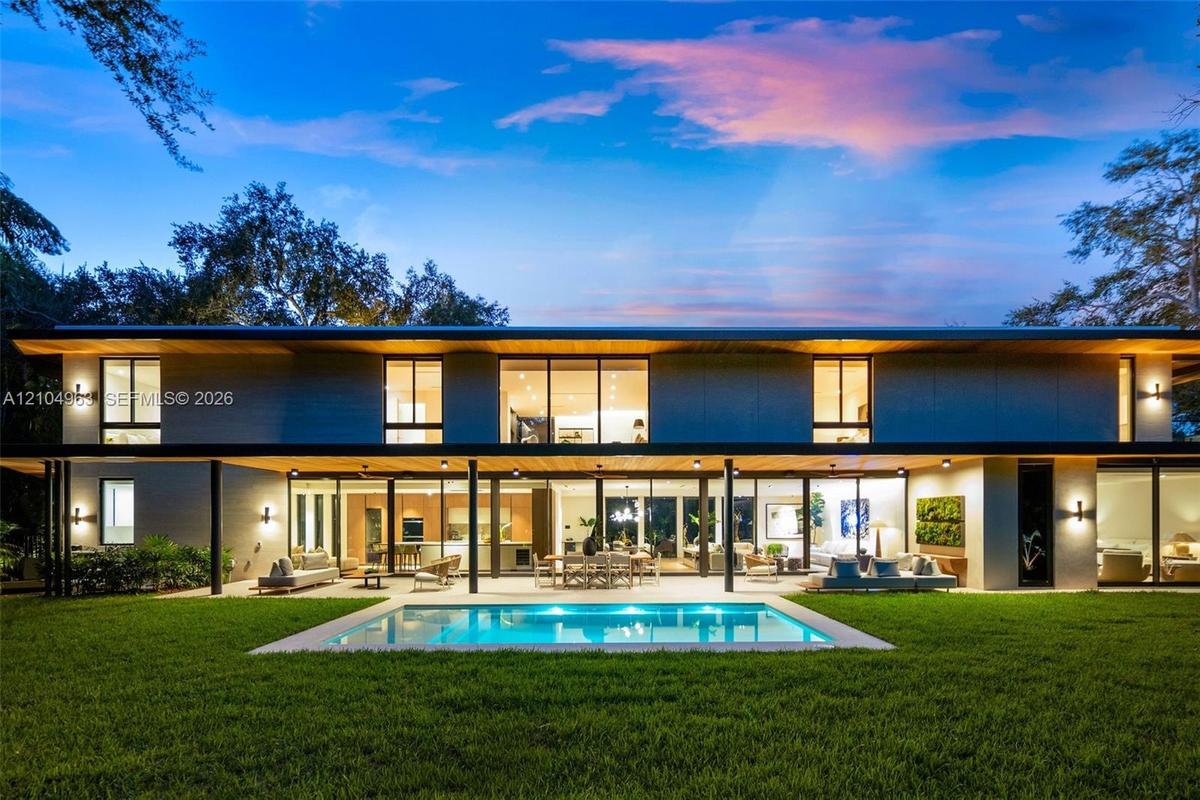Modern two-story architectural exterior featuring expansive glass walls, a covered patio with recessed lighting, a rectangular in-ground pool, and woo