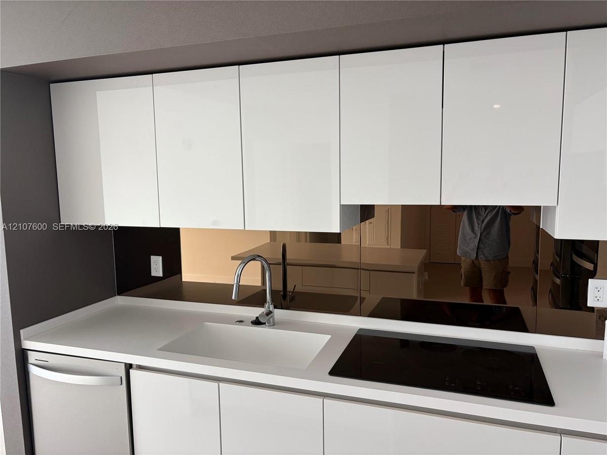 Kitchen featuring high-gloss white cabinetry, white stone-composite countertop, integrated white sink with chrome arched faucet, and a mirrored backsp