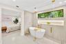 Freestanding white soaking tub enclosed by a frameless glass partition, featuring gold-tone hardware, full-height stone-finish wall tiling, a recessed