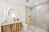 Floating wood-finish vanity with an oval vessel sink, back-lit circular mirror, walk-in shower with floor-to-ceiling tile, and brass-finish fixtures
