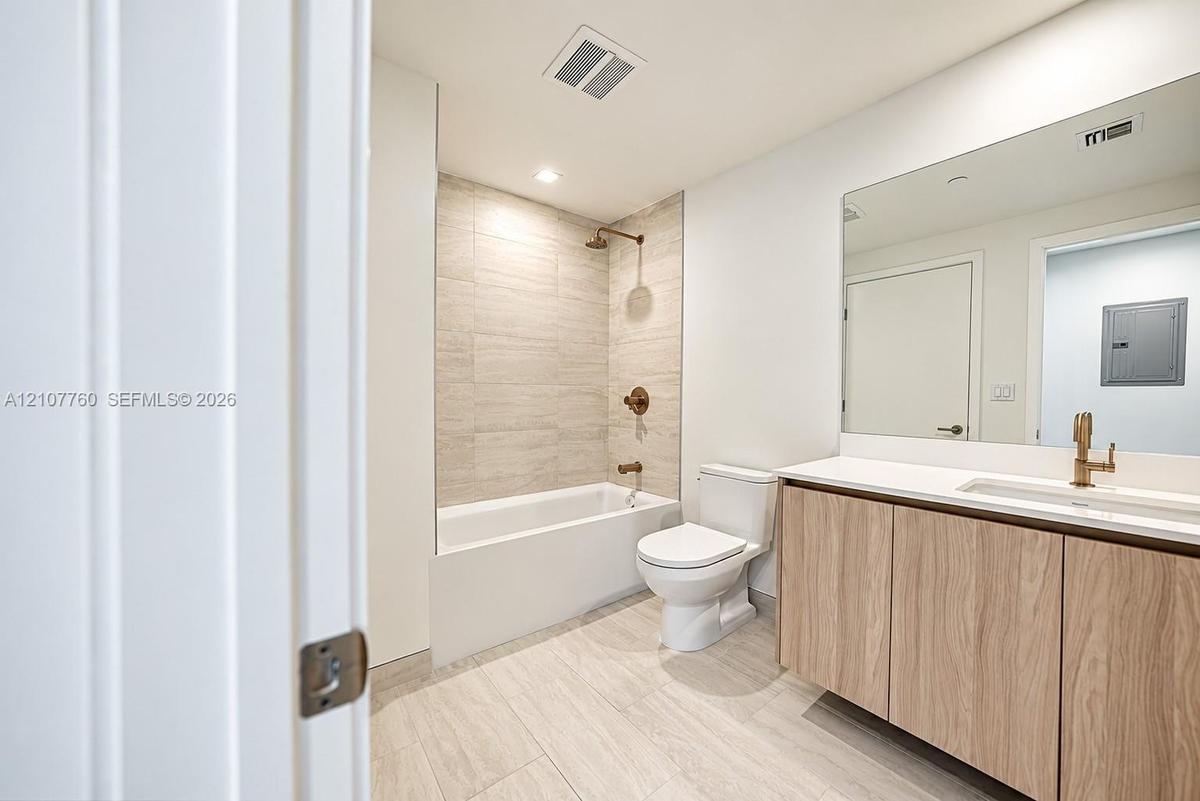 Bathroom featuring a wood-finish floating vanity, neutral-toned tile flooring, large wall-mounted mirror, and a tub with tiled surround and bronze-ton