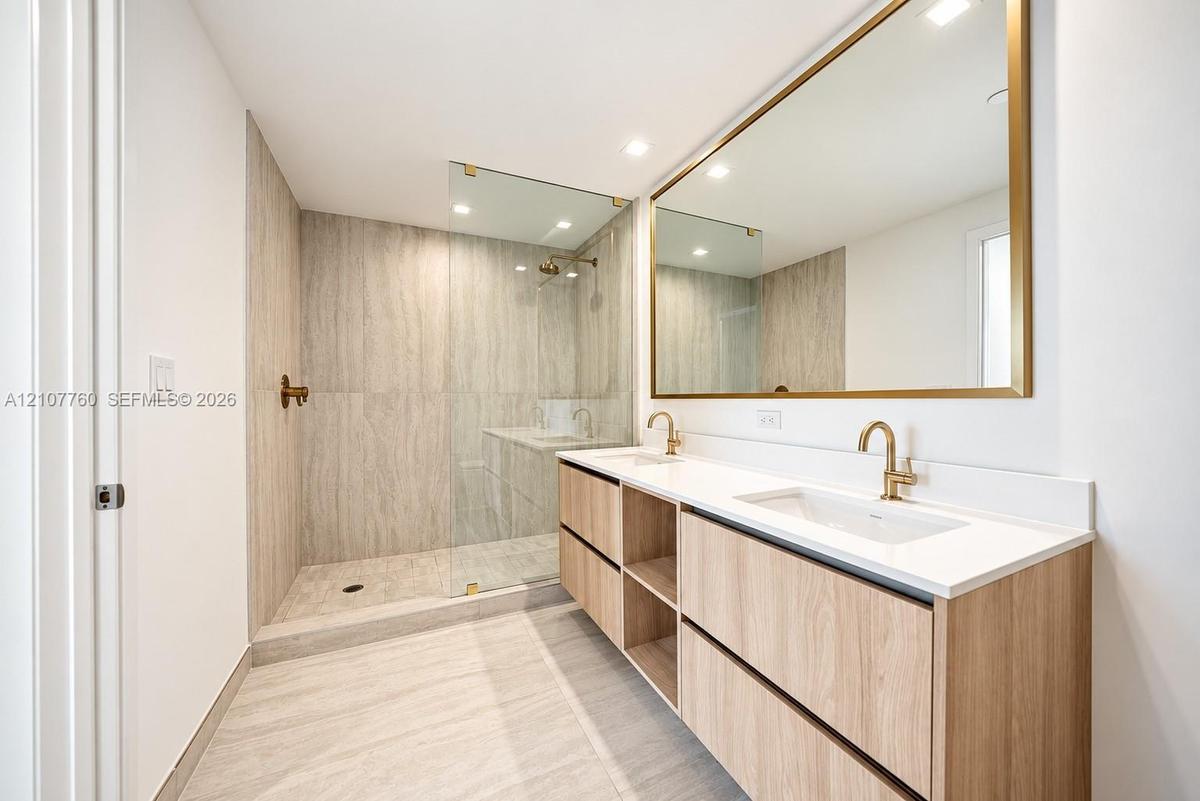 Wood-finish dual vanity with white countertop, integrated rectangular sinks, expansive gold-rimmed mirror, and walk-in shower with floor-to-ceiling st