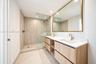 Wood-finish dual vanity with white countertop, integrated rectangular sinks, expansive gold-rimmed mirror, and walk-in shower with floor-to-ceiling st