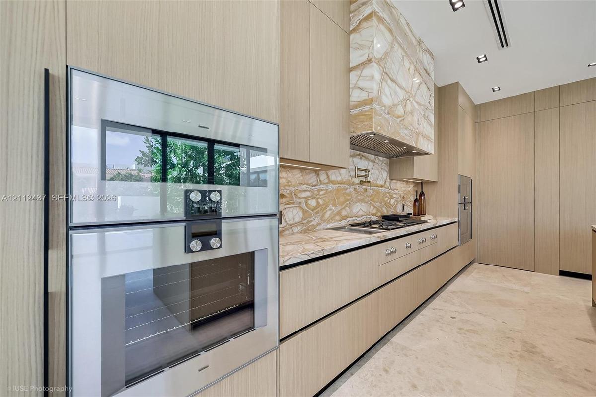 Modern kitchen featuring flat-panel wood-finish cabinetry, floor-to-ceiling stone-slab backsplash and hood surround, integrated gas cooktop with pot f