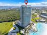 High-rise glass facade building featuring wrap-around balconies, a ground-level swimming pool, and immediate access to a man-made lagoon with a sand s