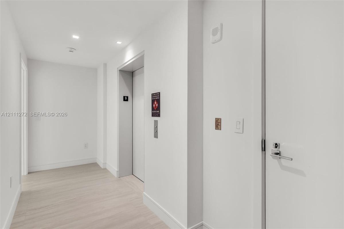 Hallway featuring light-toned wood-finish flooring, recessed ceiling lighting, and a dedicated elevator access point with a metallic call button panel