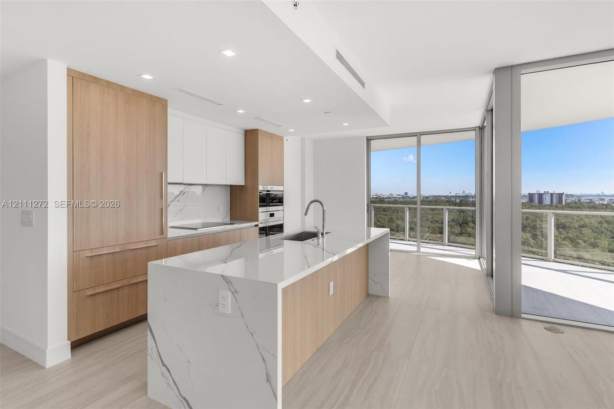 Kitchen featuring a waterfall island with a stone-finish countertop, integrated wood-finish cabinetry, built-in double wall ovens, and floor-to-ceilin
