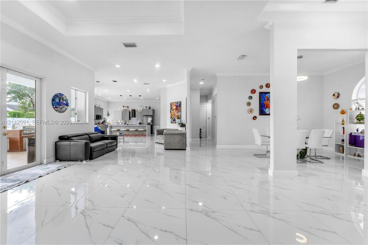 Open-concept main living area featuring large-format polished tile flooring, recessed lighting, tray ceilings, and a glass-paneled door leading to an 