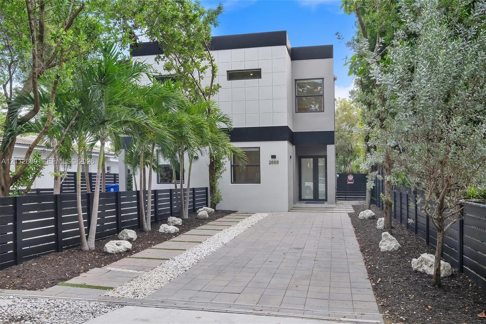 Modern two-story exterior featuring smooth stucco facade, horizontal slat privacy fencing, paved stone driveway, and curated rock landscaping