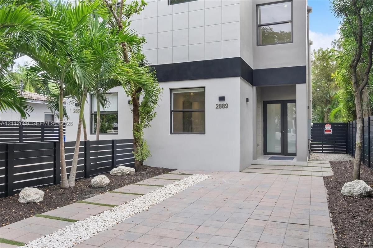 Modern two-story exterior featuring smooth stucco siding, large rectangular windows, black horizontal perimeter fencing, paver driveway, and a glass-p