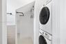Laundry area featuring stacked appliances, matte black wall-mounted metal shelving, neutral large-format floor tiling, and white wall finishes