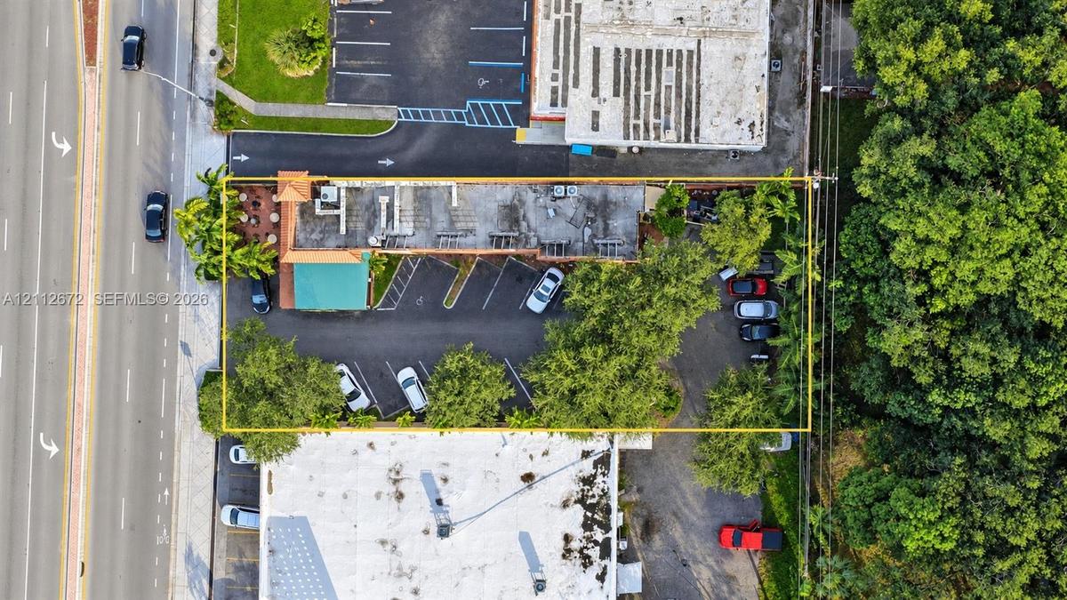 Aerial perspective of property featuring paved surface parking, mature perimeter trees, and street-front access