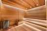 Sauna featuring light-toned wood wall and ceiling paneling, multi-level bench seating, integrated strip lighting, and a metal sauna heater