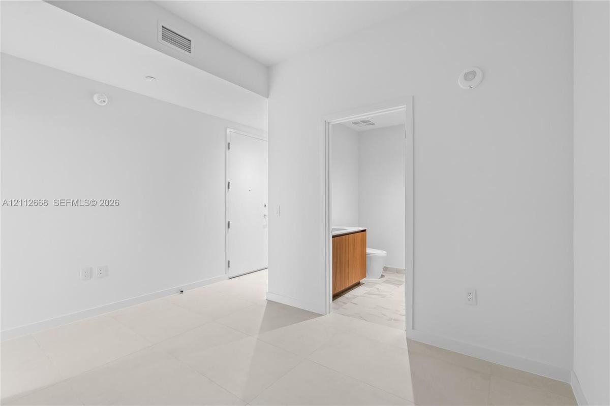 Large-format tile flooring, high ceilings, and neutral wall tones, featuring an interior doorway to a bathroom with wood-finish cabinetry and stone-lo