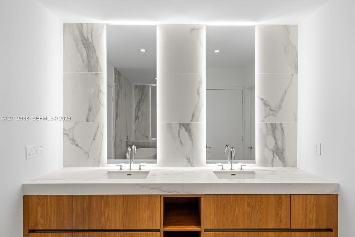Double vanity featuring a wood-finish cabinet, stone-surfaced countertop, integrated rectangular sinks, backlit vertical mirrors, and stone-tiled back