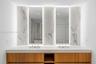 Double vanity featuring a wood-finish cabinet, stone-surfaced countertop, integrated rectangular sinks, backlit vertical mirrors, and stone-tiled back