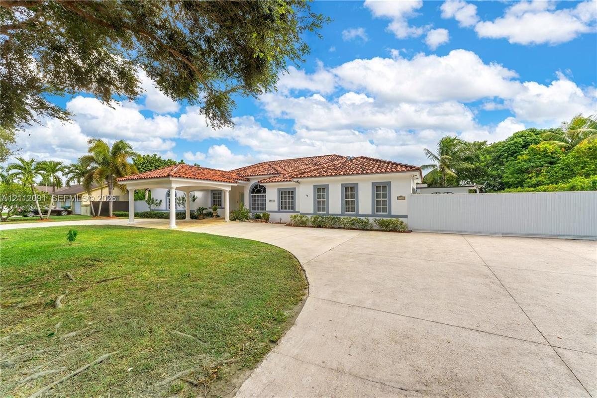 Single-story residence featuring a clay tile roof, covered porte-cochère, multi-pane windows, expansive concrete motor court, and perimeter privacy fe