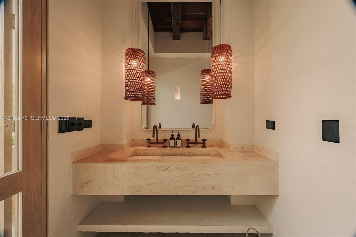 Custom stone floating vanity with an integrated basin, dual wall-mounted faucets, arched wall mirror, and exposed timber ceiling beams