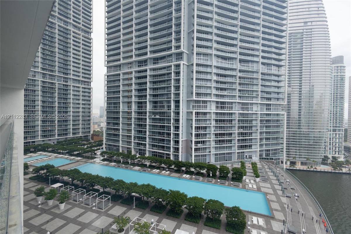 Expansive outdoor terrace featuring multi-level swimming pools, paved stone decking, landscaped trees, and high-rise tower surroundings