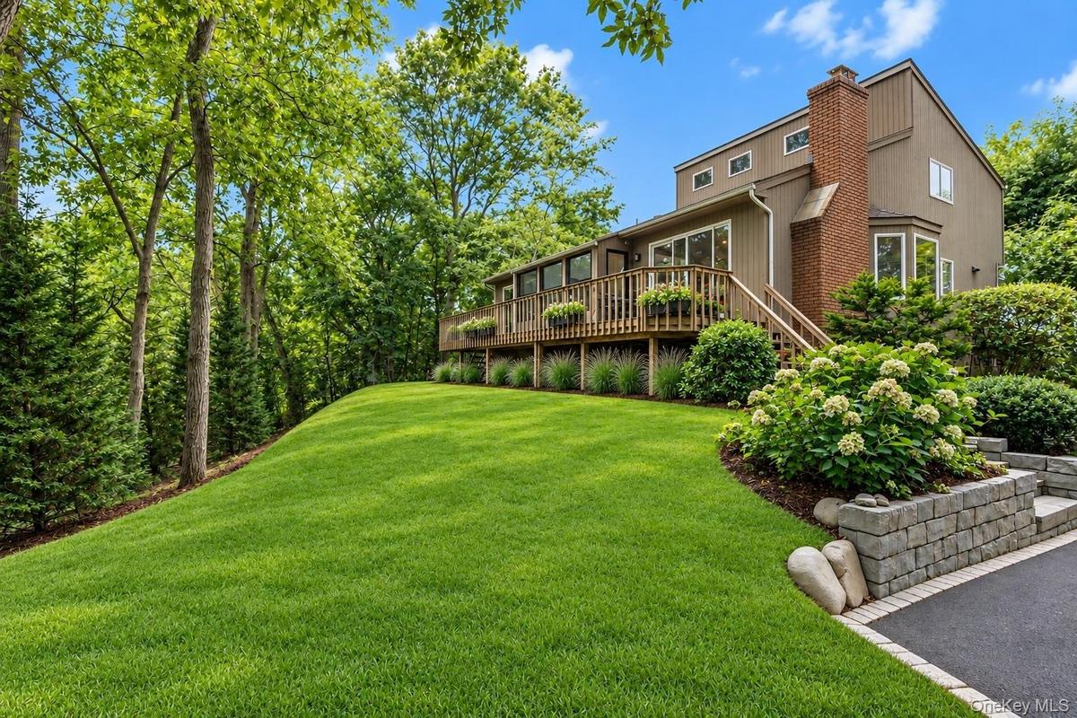 Exterior featuring vertical wood-finish siding, a red brick chimney, an elevated wood deck, stone-block retaining walls, and a manicured lawn