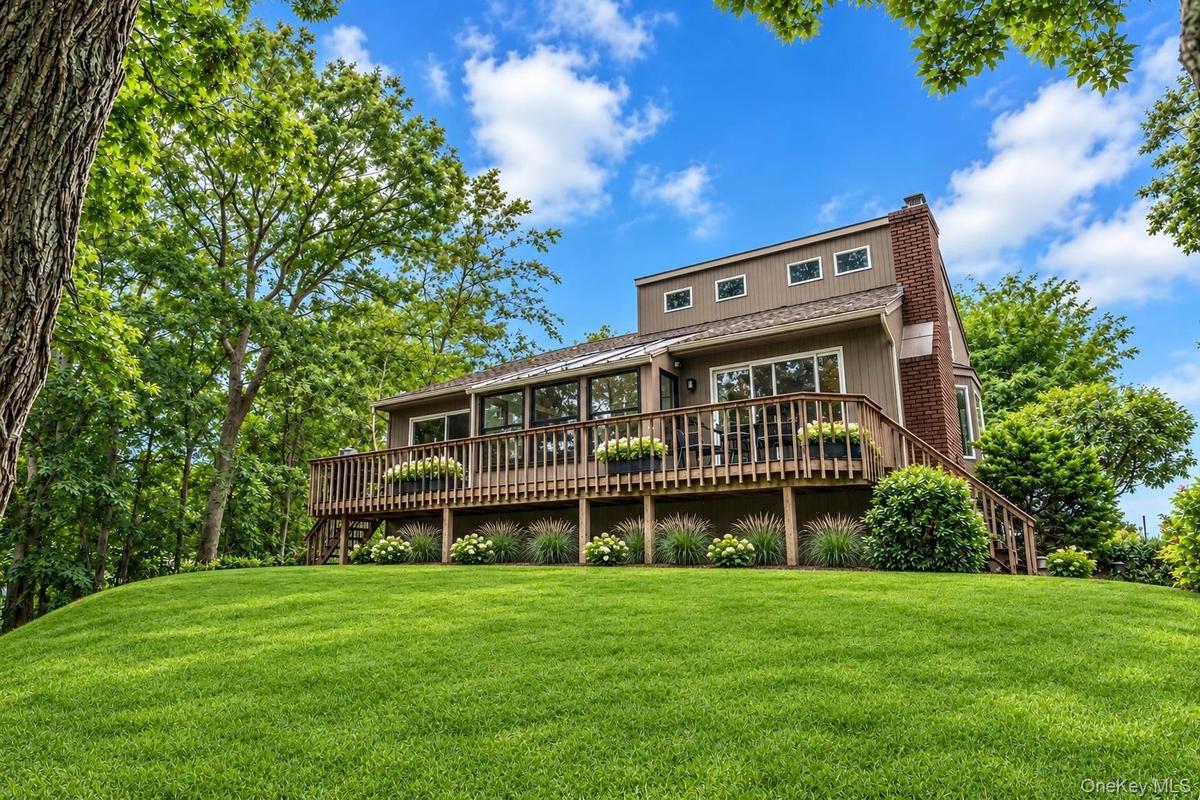 Exterior featuring a wrap-around wooden deck, brick chimney, horizontal wood siding, and expansive manicured lawn
