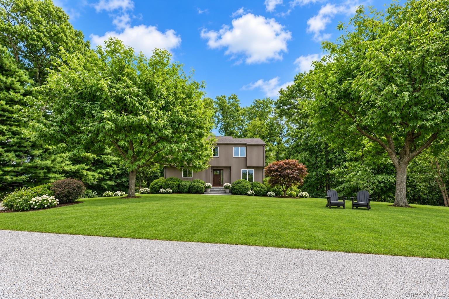 Two-story residence featuring wood-tone exterior cladding, expansive manicured lawn, mature shade trees, and a crushed stone driveway