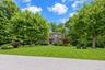Two-story residence featuring wood-tone exterior cladding, expansive manicured lawn, mature shade trees, and a crushed stone driveway