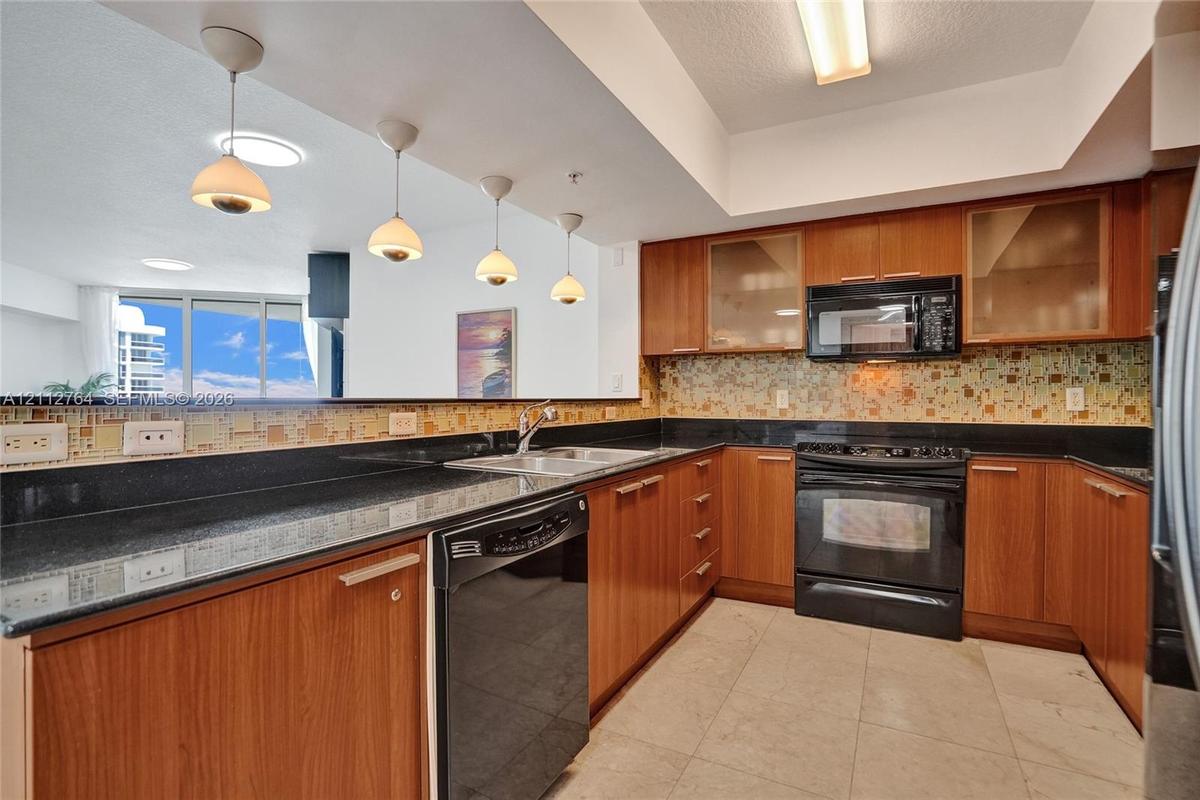 Kitchen featuring natural wood-finish cabinetry, dark solid-surface countertops, multi-toned glass tile backsplash, tile flooring, and four pendant li
