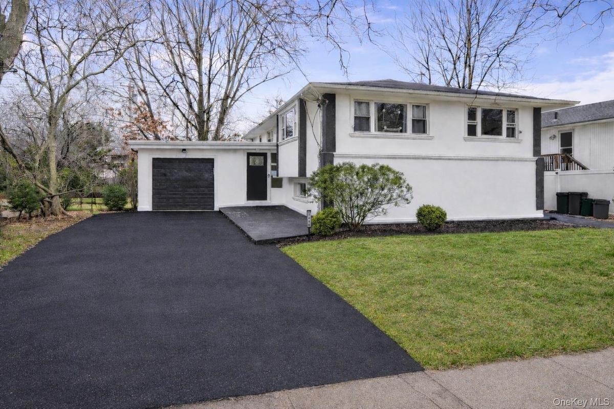 Split-level exterior featuring a light-toned smooth finish, dark-trimmed accents, an asphalt driveway, single-car garage, and landscape greenery