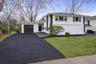 Split-level exterior featuring a light-toned smooth finish, dark-trimmed accents, an asphalt driveway, single-car garage, and landscape greenery