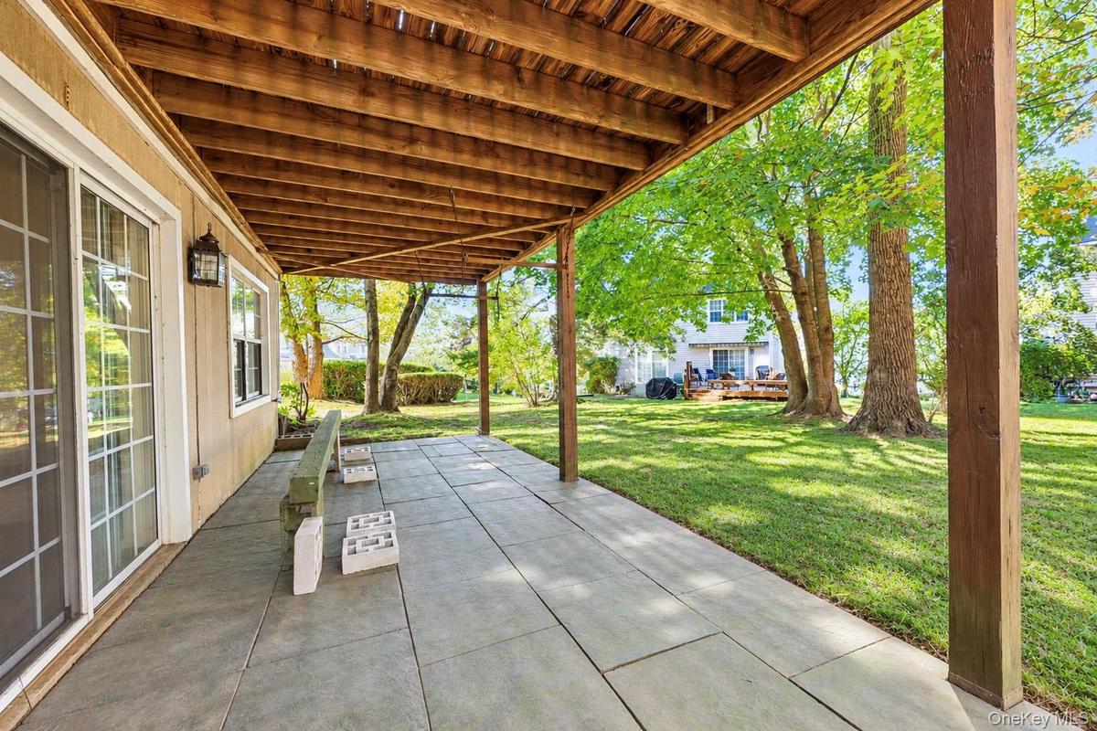 Covered patio featuring large-format stone-style pavers, exposed wood ceiling beams, sliding glass doors, and direct access to a lawn with mature tree