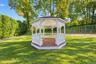 Octagonal gazebo featuring a white wood frame, integrated wrap-around seating, and wood-finish decking, situated on a manicured lawn with mature trees