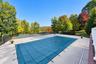 Outdoor area featuring a swimming pool with a mesh safety cover, expansive concrete pool deck, fenced tennis court, and mature trees