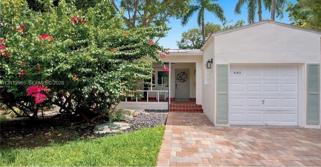 Single-story exterior featuring a paver driveway, attached garage, covered front porch, and mature landscaping