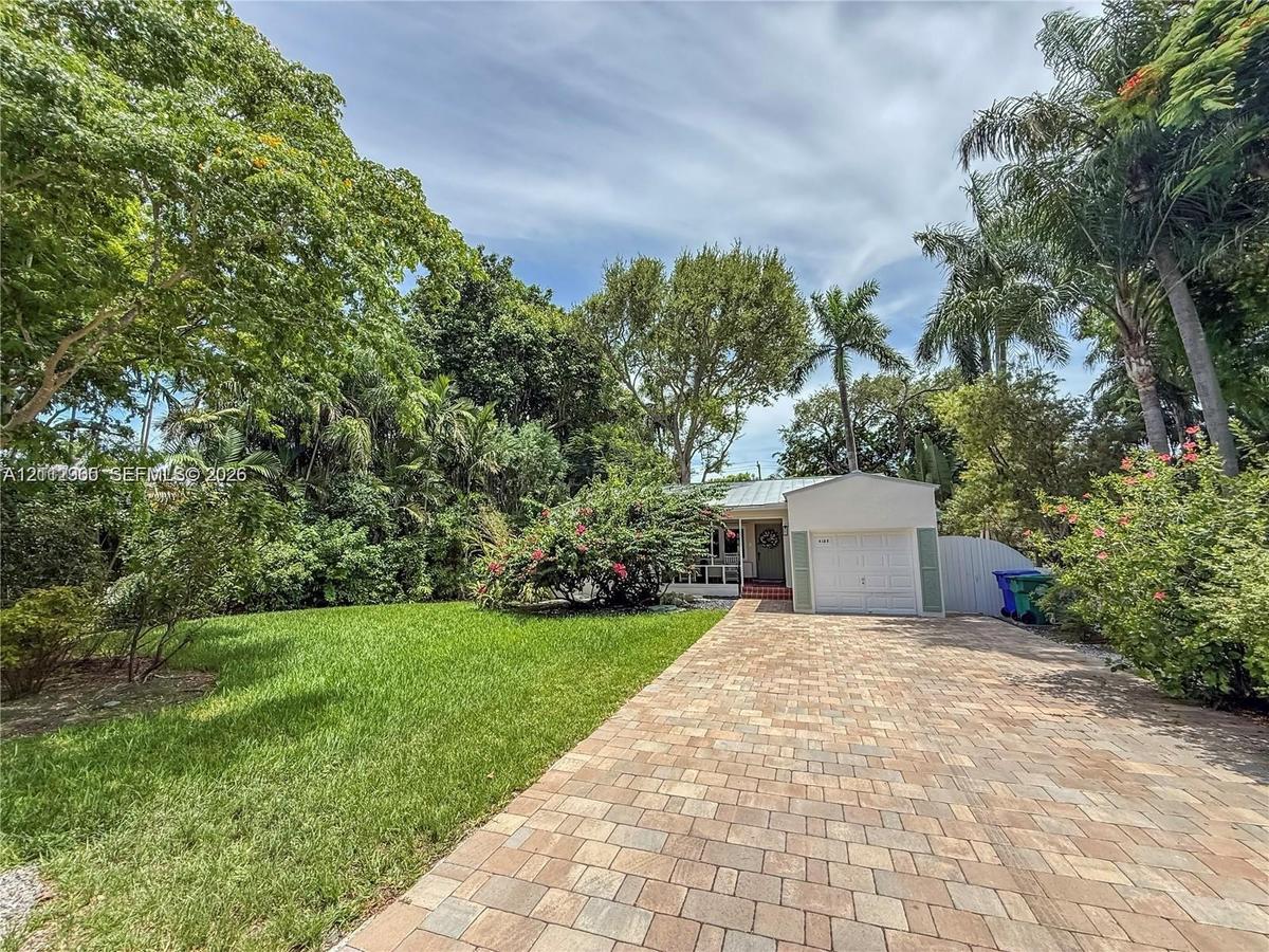 Paver driveway leading to a single-car garage, complemented by extensive mature landscaping and a lawn area