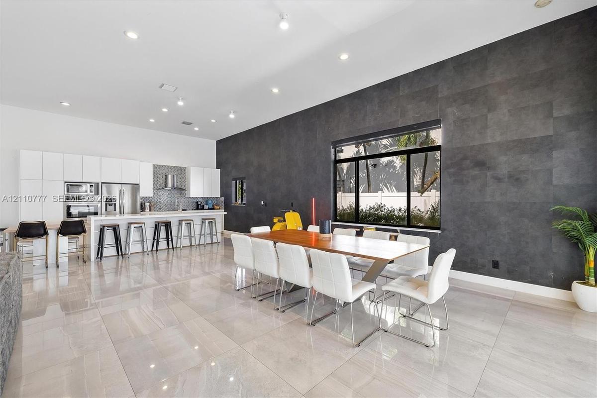 Modern kitchen and dining area featuring high-gloss white cabinetry, expansive stone tile flooring, a dark charcoal textured accent wall, recessed lig