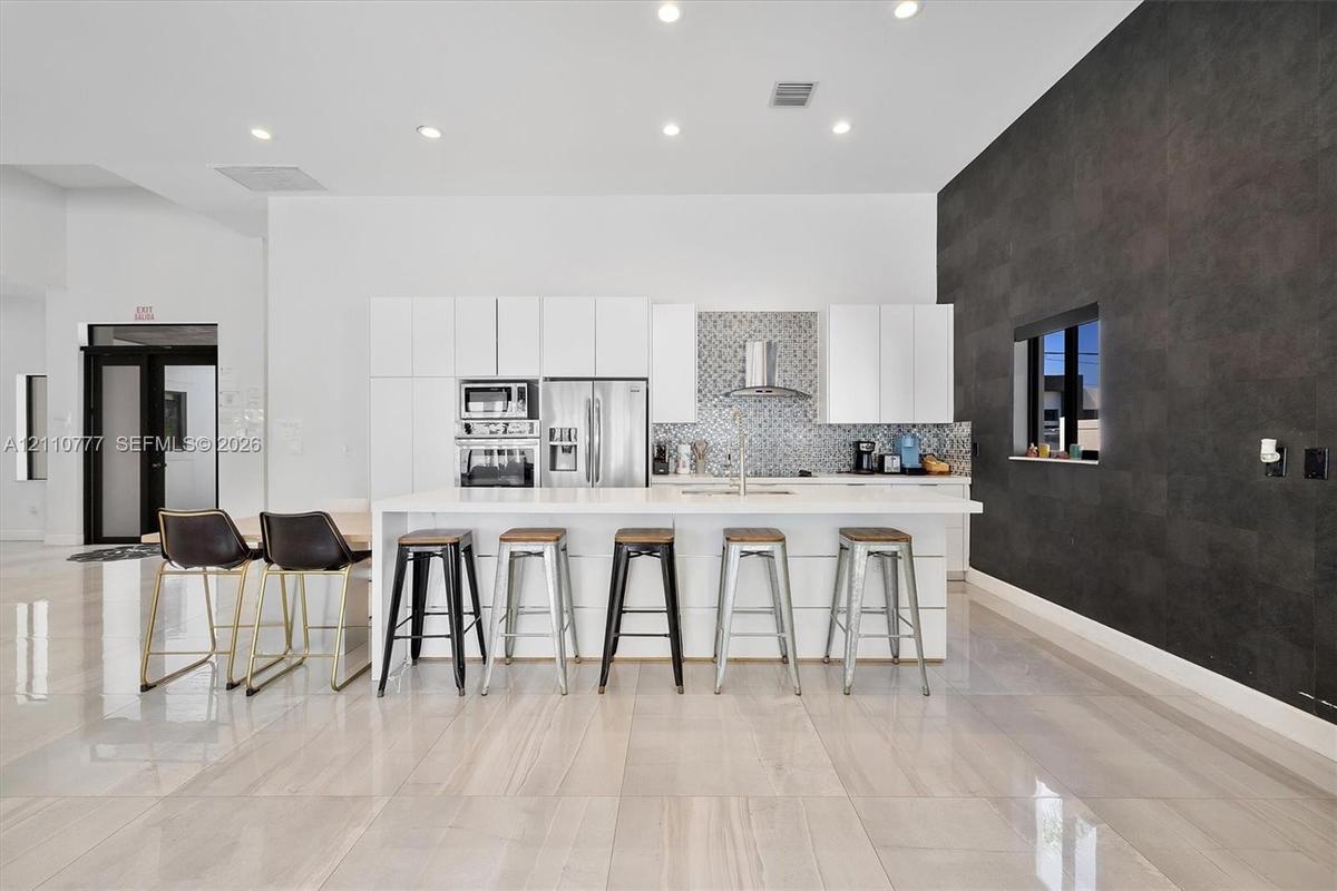 Modern kitchen featuring flat-panel white cabinetry, a waterfall-edge kitchen island, stone-finish tile flooring, a mosaic tile backsplash, and a dark