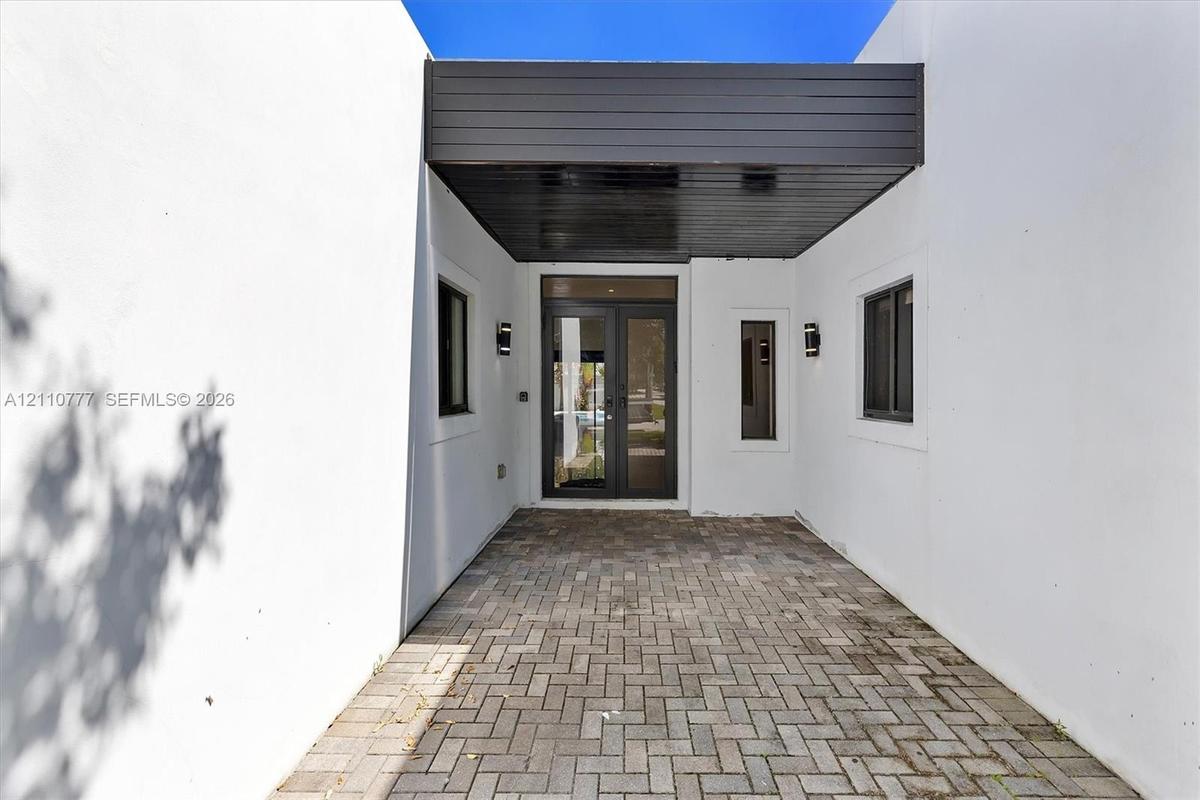 Covered entryway featuring a herringbone-patterned paver floor, minimalist smooth-stucco exterior walls, dark wood-finish ceiling paneling, glass-pane