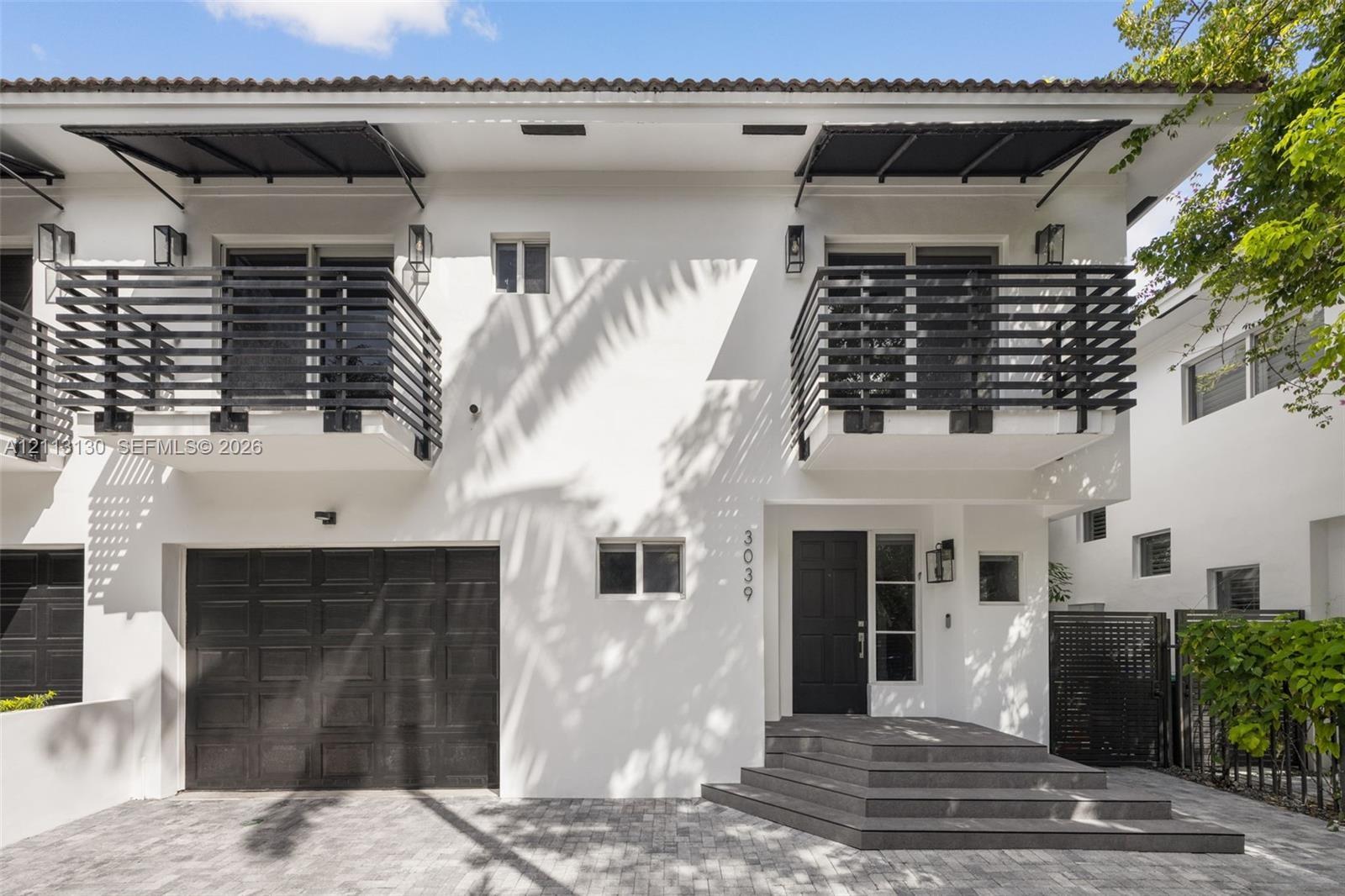 Two-story stucco exterior featuring horizontal black metal balcony railings, a multi-paneled garage door, geometric entry steps, and matching black me