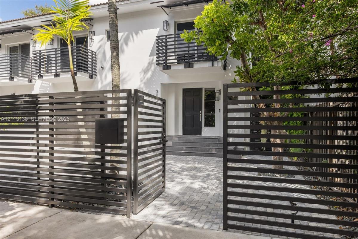 Exterior facade featuring a paved stone driveway, horizontal slat metal privacy fencing, solid panel entry door, and upper-level balconies with matchi