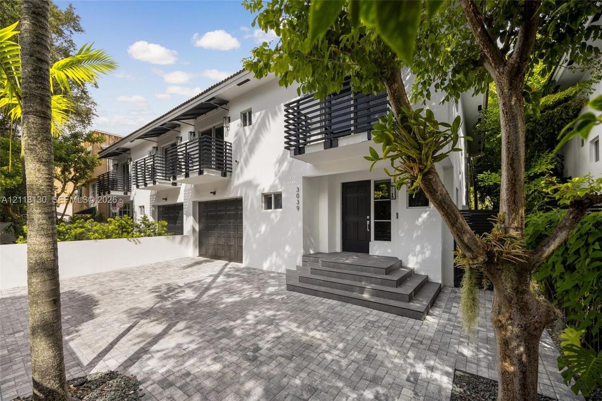 Stucco exterior featuring dual-level black metal balconies, a paved stone driveway, an attached garage, and tiered concrete front entry steps