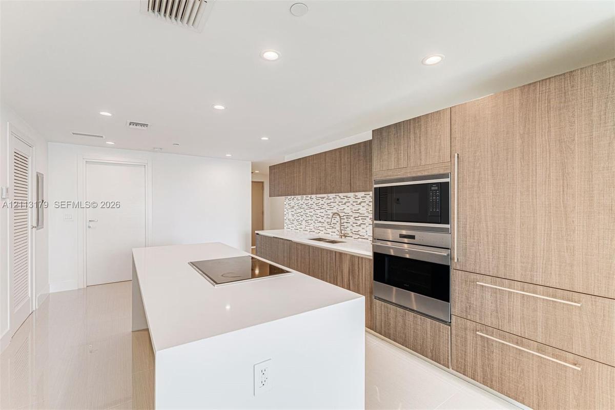Kitchen featuring wood-finish cabinetry, a white countertop island with an integrated cooktop, stainless steel wall-mounted oven and microwave, stone 