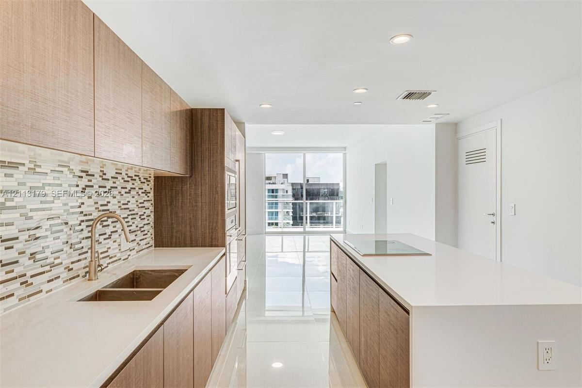 Kitchen featuring wood-finish cabinetry, stone-surface countertops, mosaic tile backsplash, integrated stainless steel appliances, and a central islan