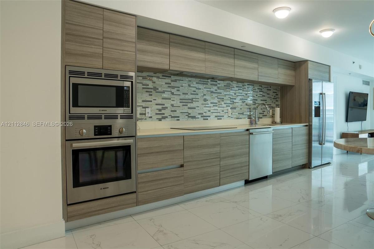 Kitchen featuring flat-panel wood-finish cabinetry, stainless steel appliances, multi-tonal glass tile backsplash, stone-finish countertops, and polis