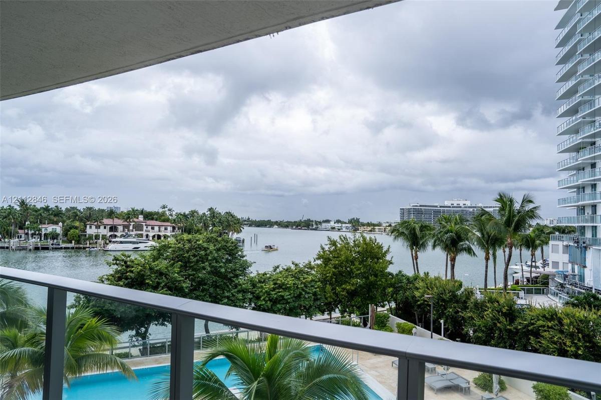 Covered balcony with a metal railing, overlooking an outdoor swimming pool, lush landscaping, and a waterfront setting