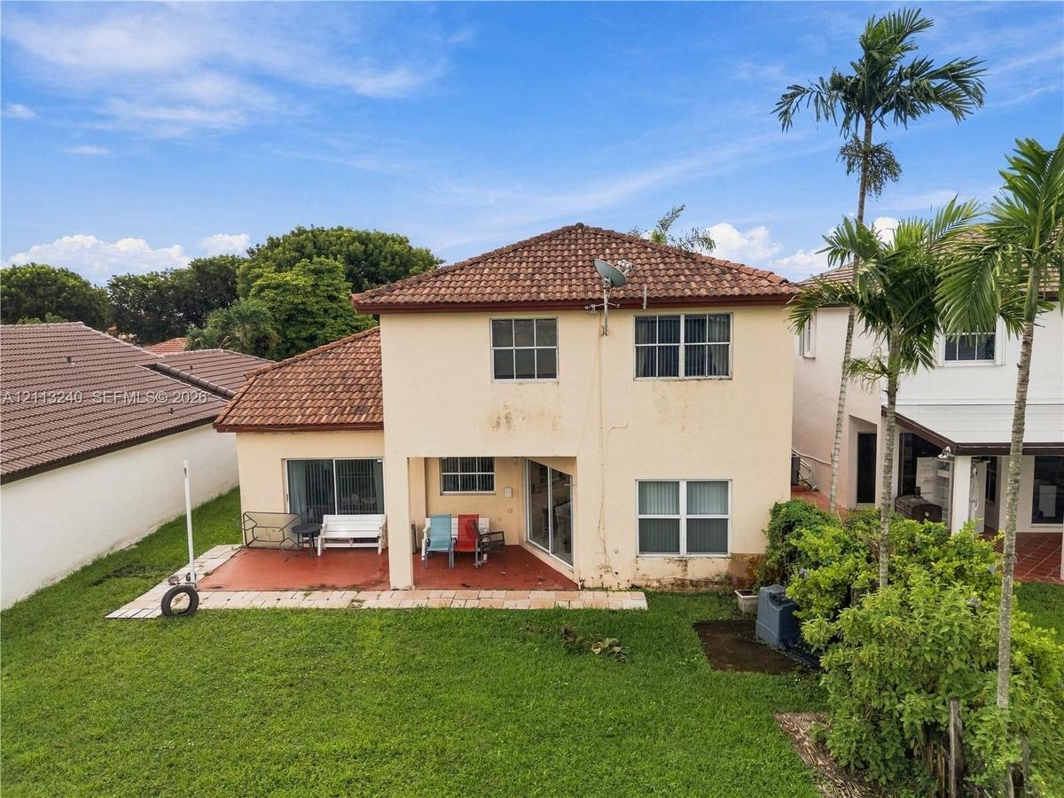 Two-story exterior featuring a clay tile roof, stucco facade, covered rear patio, and expansive lawn with mature palm trees