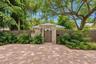 Paver driveway featuring a stone-clad arched entryway with a wood-finished privacy gate, surrounded by mature trees and lush landscaping