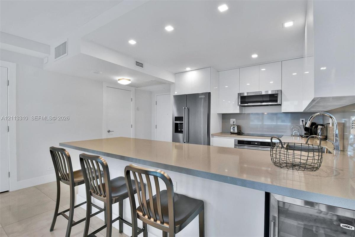Kitchen featuring high-gloss white cabinetry, a neutral stone-surface peninsula, stainless steel appliances, recessed ceiling lighting, and tile floor