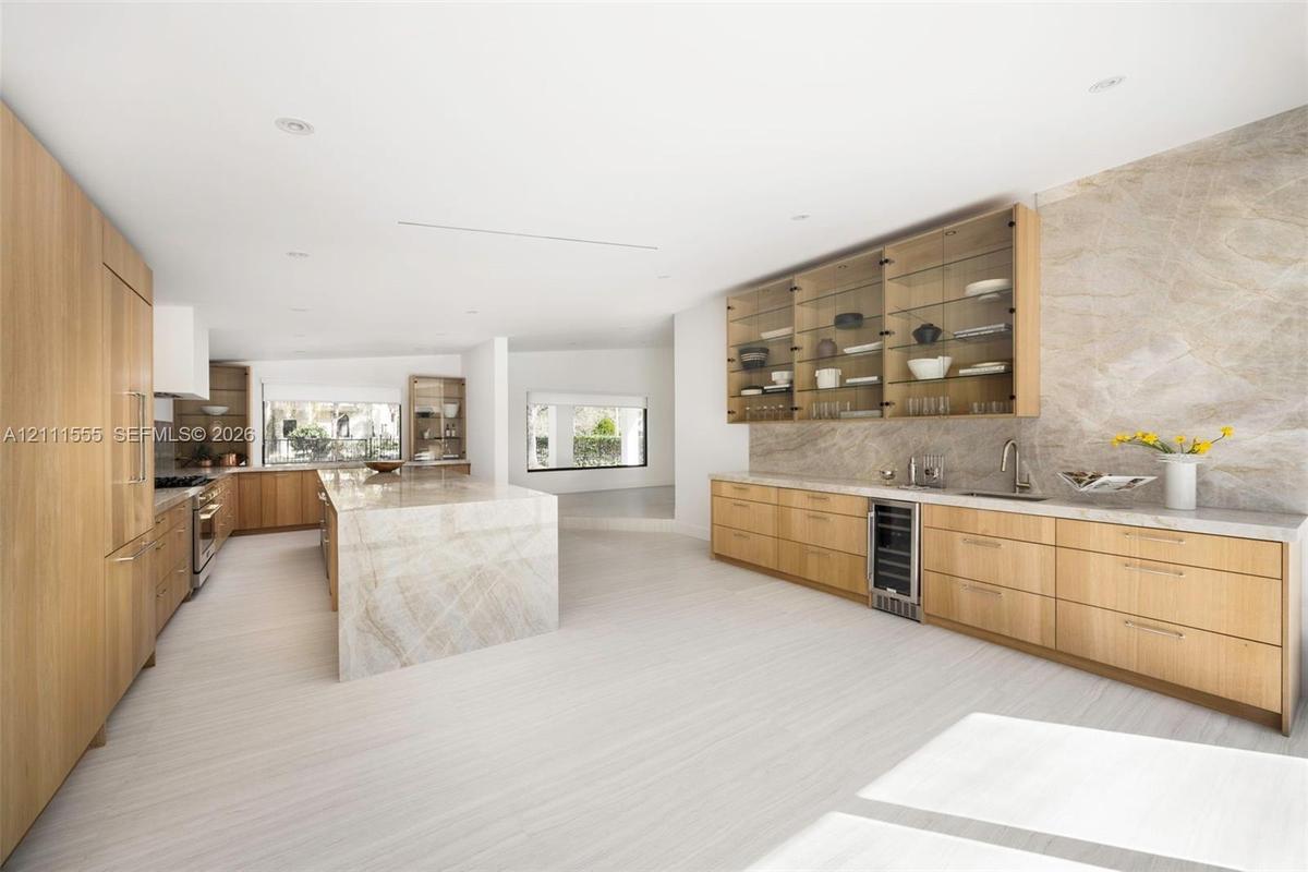 Custom wood-finish cabinetry, expansive stone-clad island, full-height stone backsplash, glass-front upper storage, and integrated beverage cooler