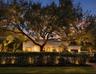 Single-story residence featuring light-colored exterior finish, mature specimen trees, perimeter metal fencing, and integrated outdoor lighting